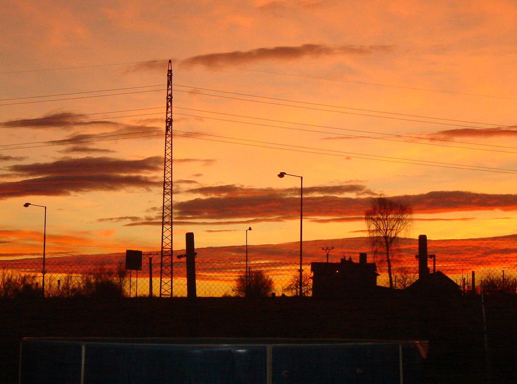 Fotografie zimního západu slunce - celá obloha je oranžová s červenými mraky, naproti nim vystupují černé siluety okolního domku a stromů a pouličních lamp.