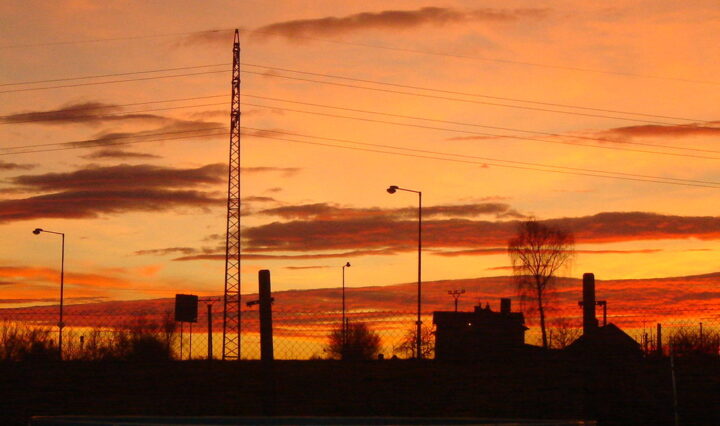 Fotografie zimního západu slunce - celá obloha je oranžová s červenými mraky, naproti nim vystupují černé siluety okolního domku a stromů a pouličních lamp.